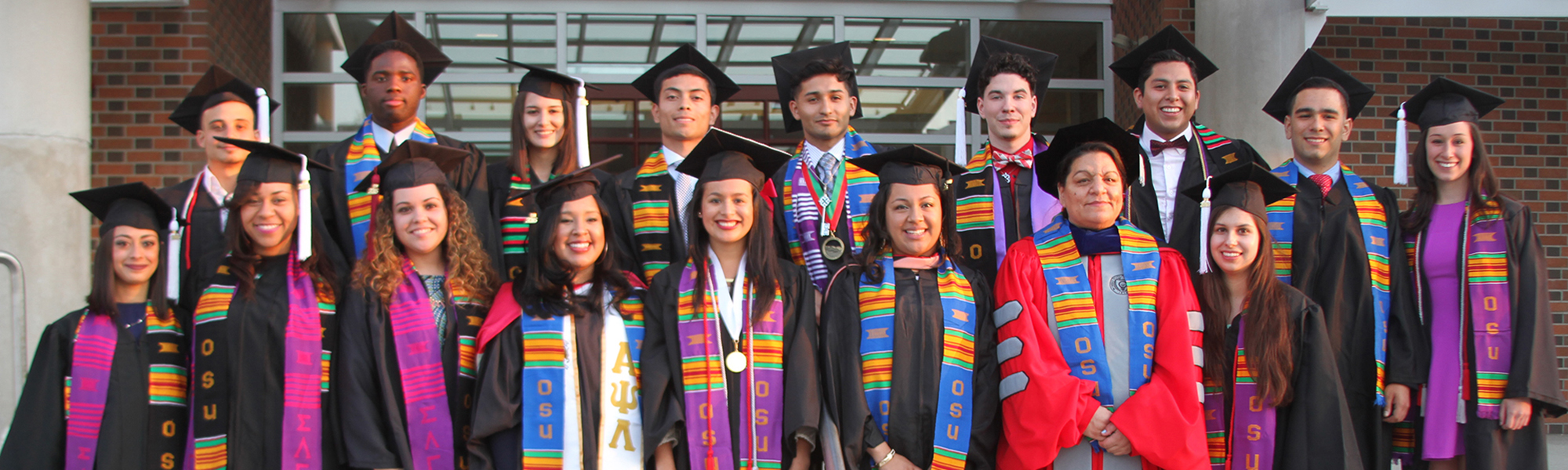 Latine students in cap and gown and PhD hood stand on seps with stoles around their necks