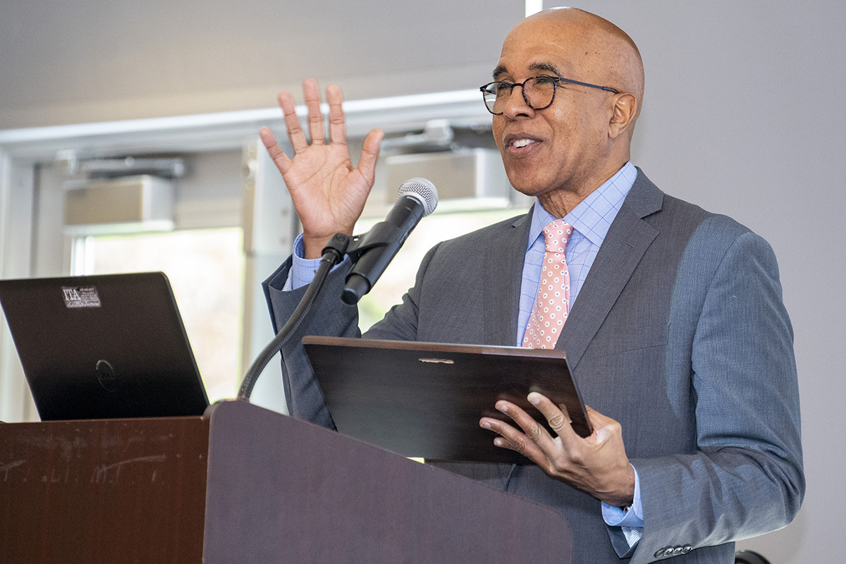 Man in a grey suit stands at a podium and raises one hand