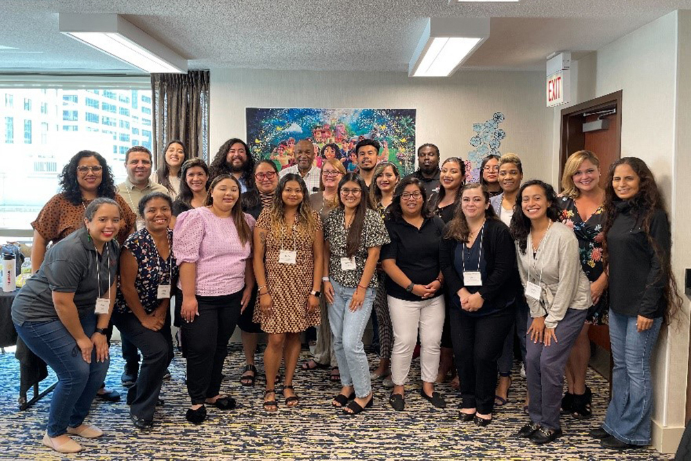 group of Latine students in business casual dress stand in front of a colorsful painting.