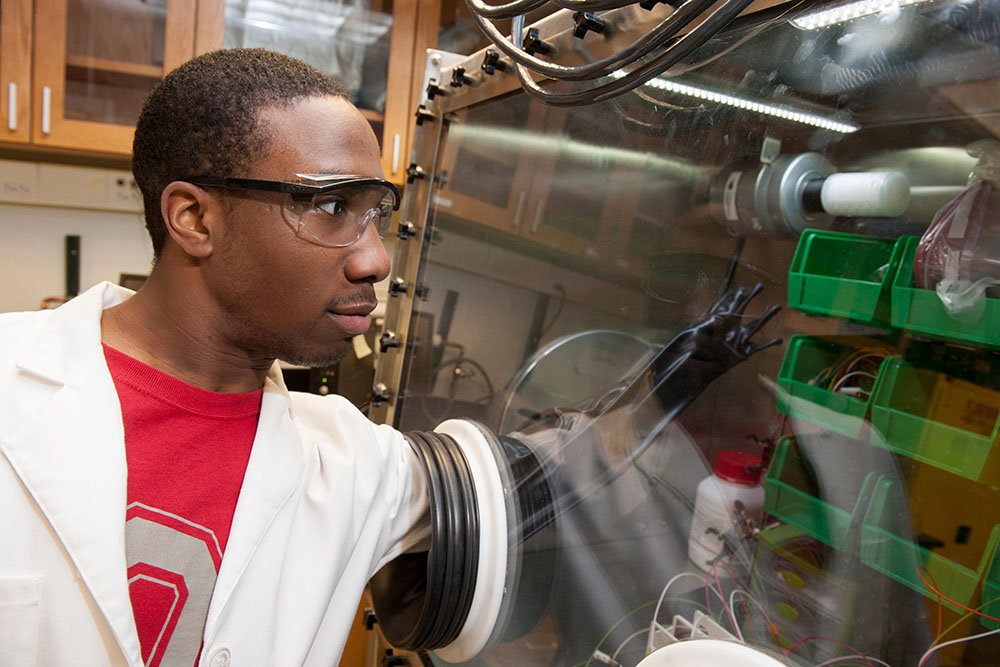 A student in goggle and a lab coat reaches through a glove in a glass panel