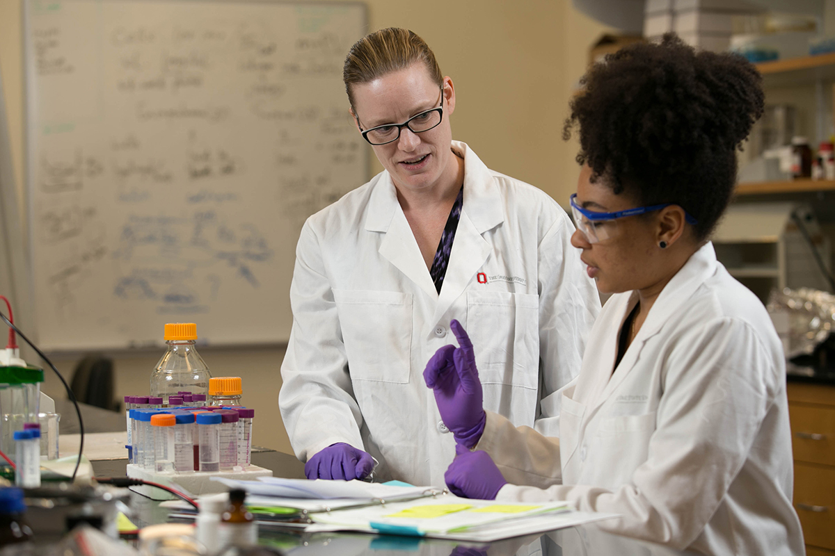 Two women in lab coats work together with lab equipment