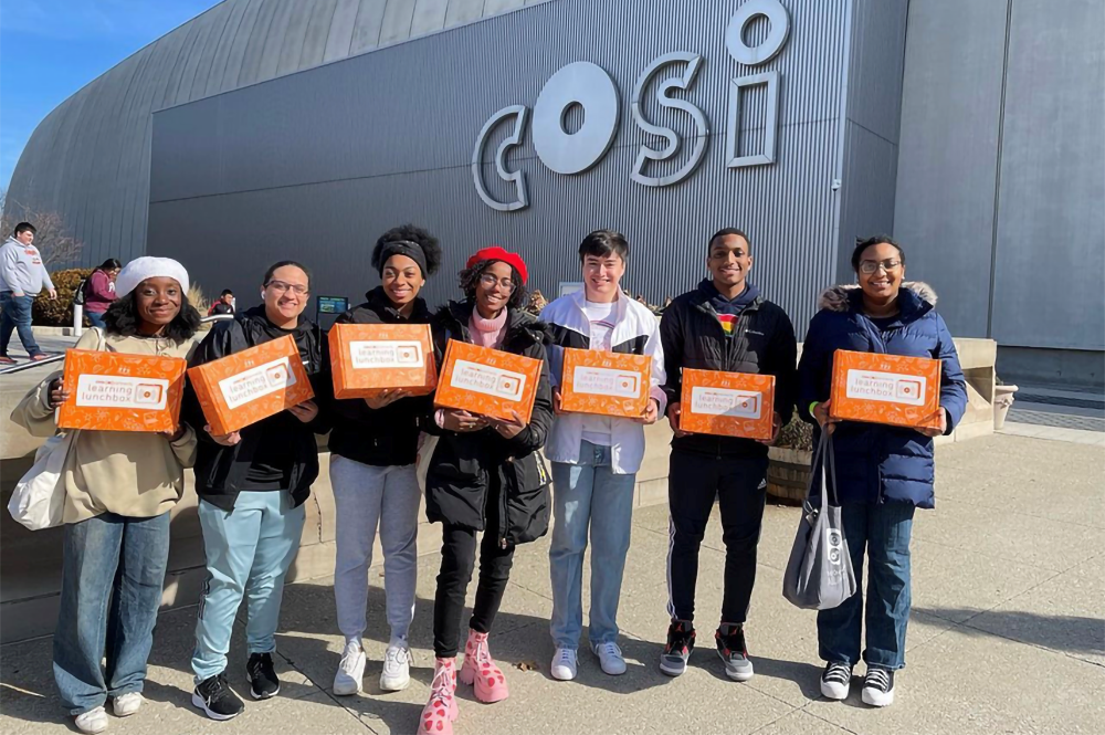 Seven students hold orange boxes outside of COSI