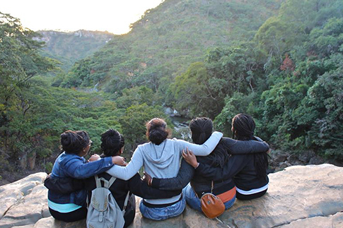 the back of 5 students sitting on a rock ledge with their arms around each other looking at moutains