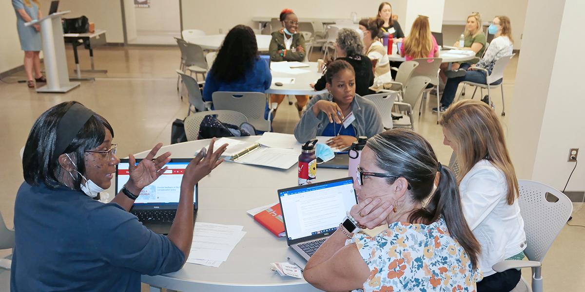 Diverse women sit around several tables with laptops and notebooks, talking