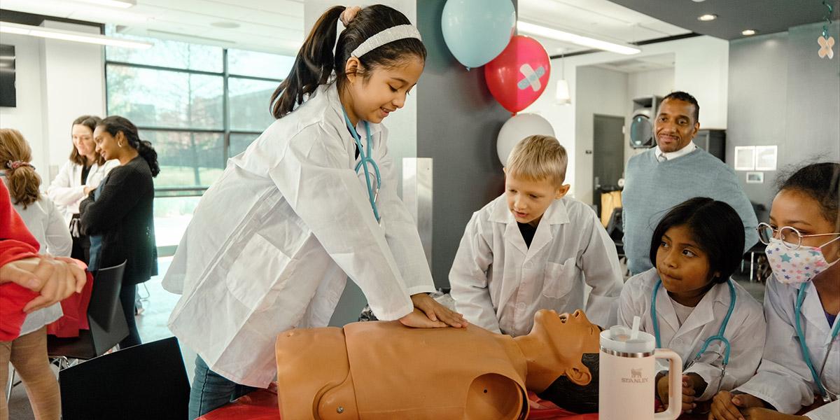 Young student in white lab coat performs CPR on a dummy while other students look on