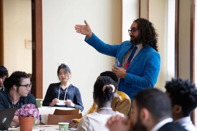 A man in a blue suit gestures to students sitting around a table