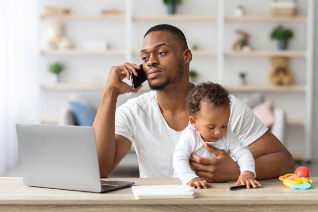 Man talking on a cell phone while holding a baby who is playing with toys on a table with a laptop