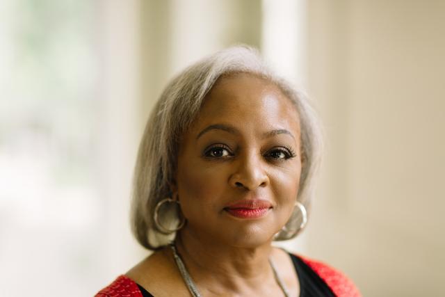 Carol Anderson in an orange cardigan and a black shirt with silver hair and large silver earrings