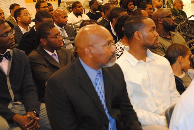 Black men sitting in chairs listen to a speaker