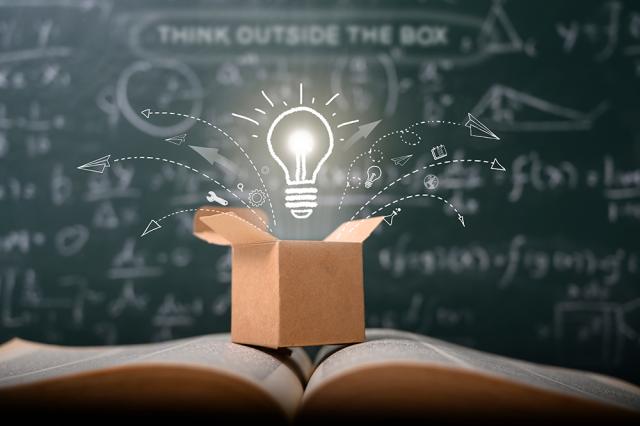 Lit lightbulb above a box sitting on an open book with scholarly equations on a chalkboard behind it.