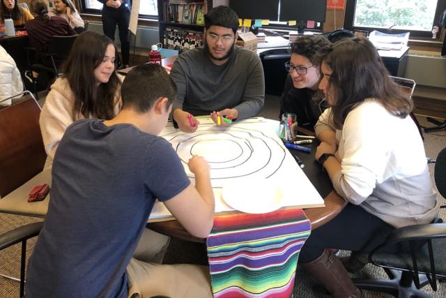 Students sit around a table with a colorful Mexican rug on it, using colored markers