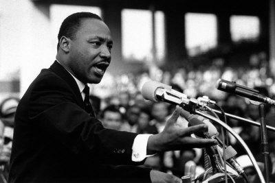 MLK in a black suit speaking at a podium with a lot a microphones