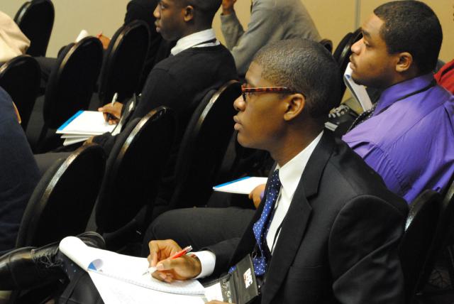 Black man with glasses listens to a speaker and takes notes