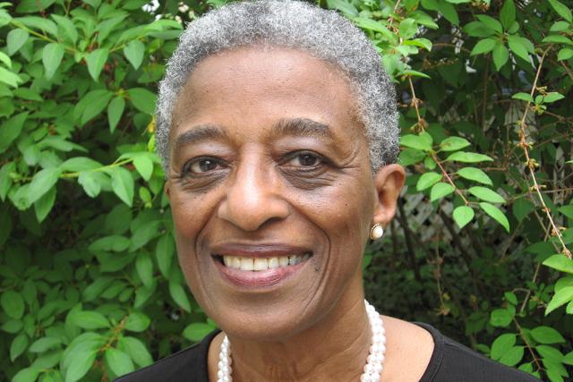 Rudine Sims Bishop, a Black woman with short grey hair, in a black dress with a pearl necklace in front of greenery