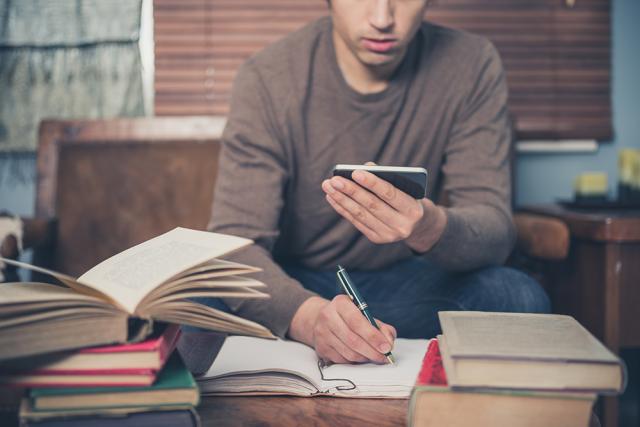 White male student with open books and phone, taking notes