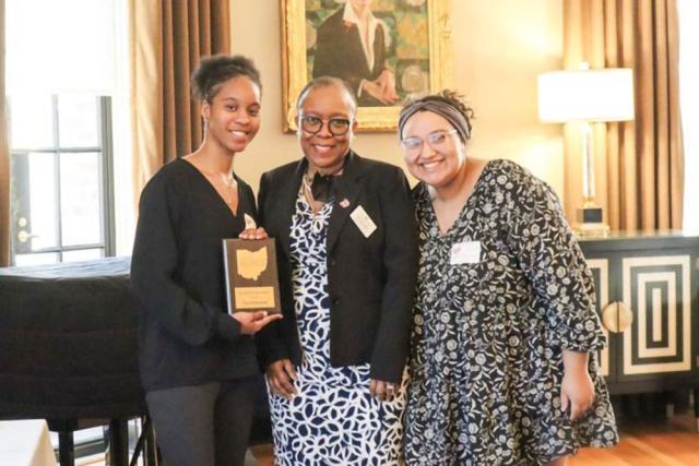 Three women stand together with the woman on the left holding an award