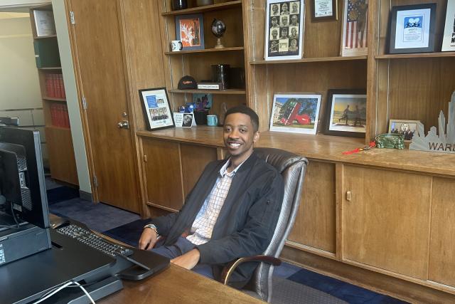 Benjamin Brooks, a Black male, sits smiling at an office desk with photos behind him