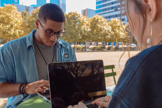 Two students sit outside at a table with laptops