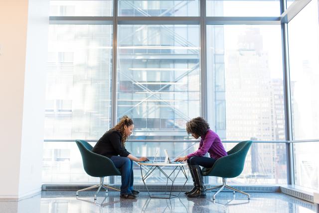 Two people sitting in front of a large window in an office building, working on laptops at a table